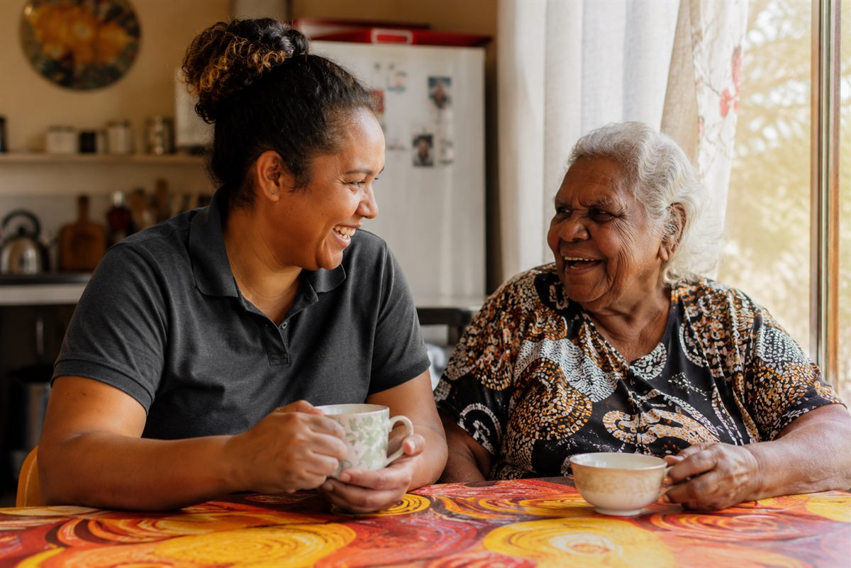 A support worker and an older Aboriginal woman sharing tea and laughing at a kitchen table